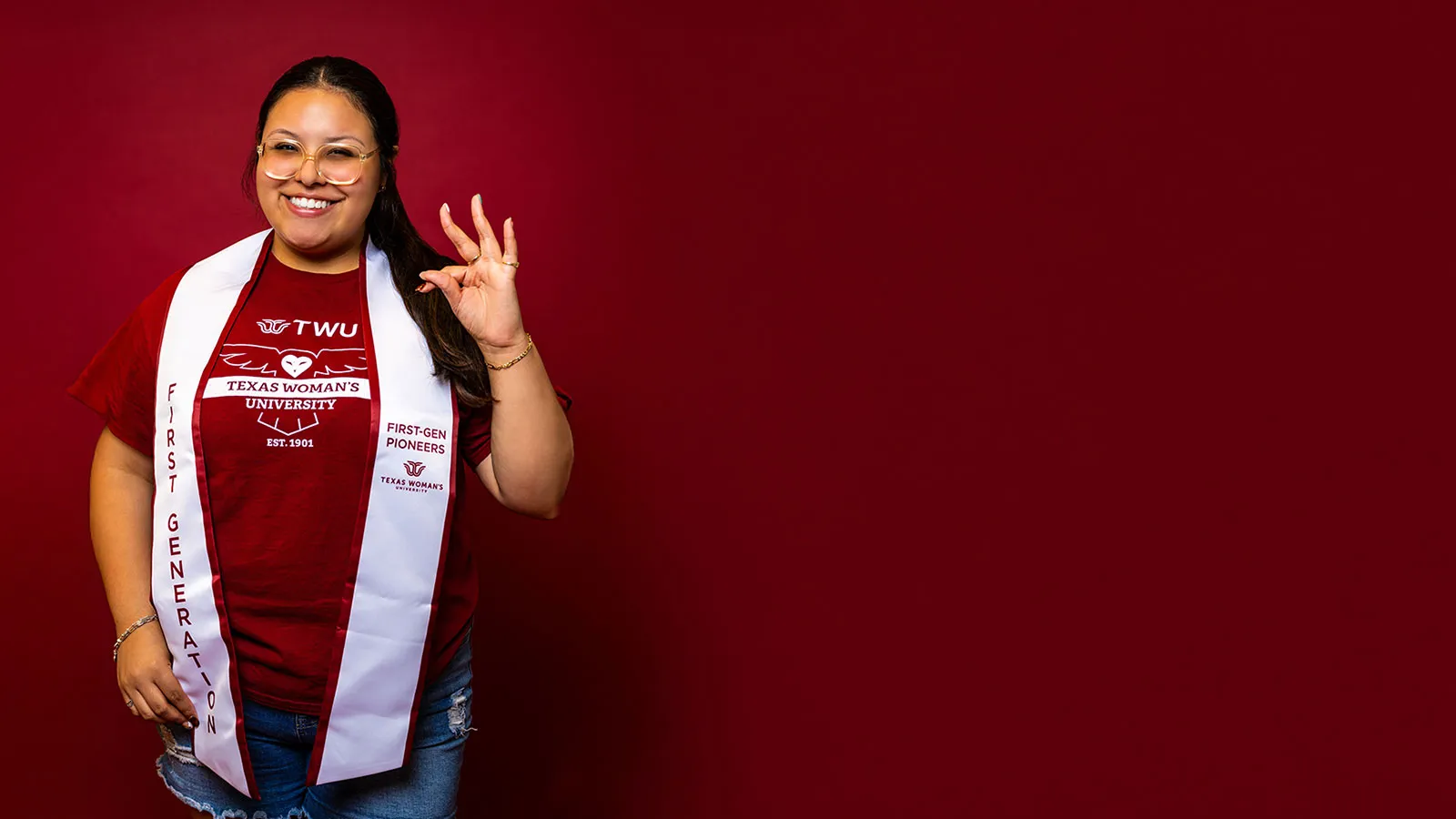 ̽¥ student Celeste Aldaco makes the ̽¥ hand sign while wearing a maroon and white sash that says 'First-Gen Pioneers'