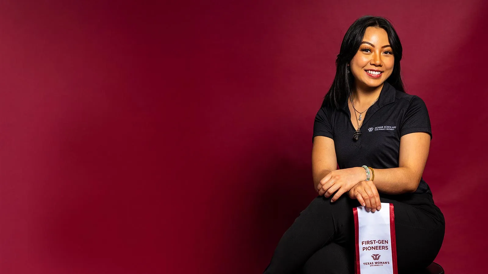 A ̽��¥ student dressed in black and holding up a white ribbon with the words 'First Generation' in maroon letters