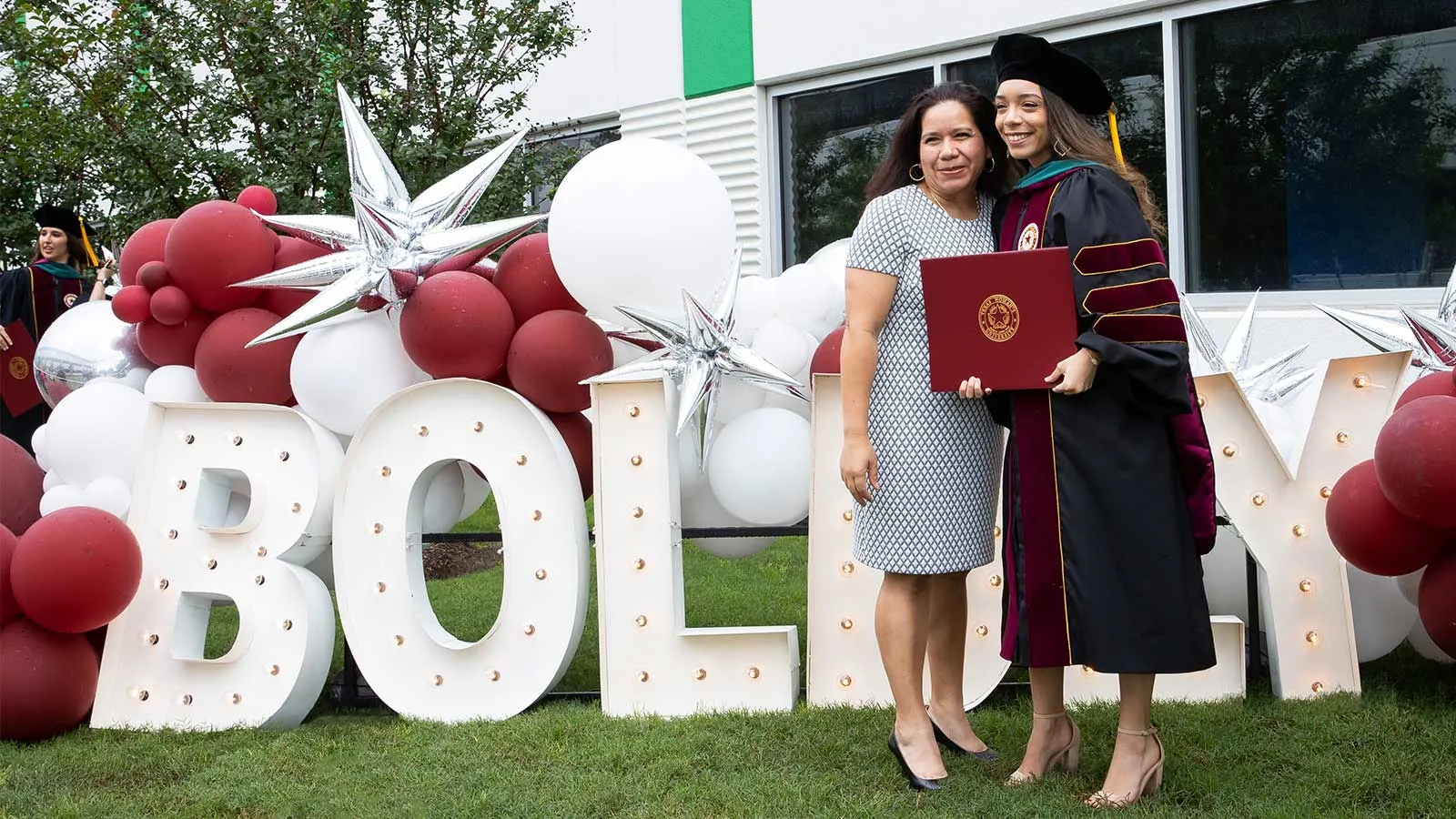 A TWU student and parent posing with a diploma in front of a sign that reads Boldly