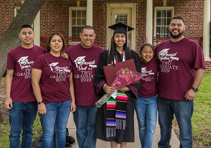 A TWU gradaute dressed in academic regalia with her family all wearing matching maroon t-shirts.