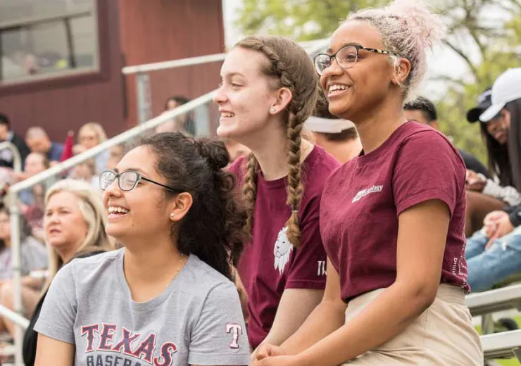 Three TWU first-year students sit in the stands and cheer during a softball game.