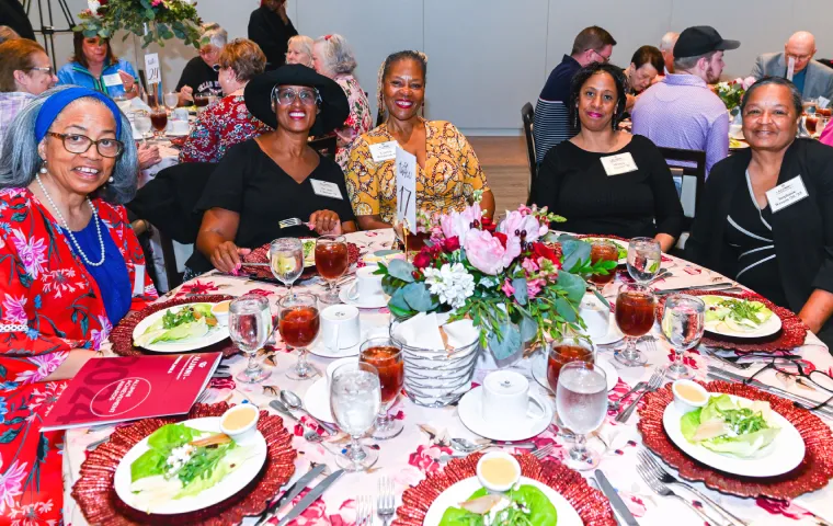 A group of alumni sit around a plated meal at a table set with flowers. They're all smiling at the camera.