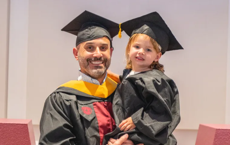A man smiles and holds up his daughter at the TWU Family Graduation ceremony. They both wear graduation regalia.
