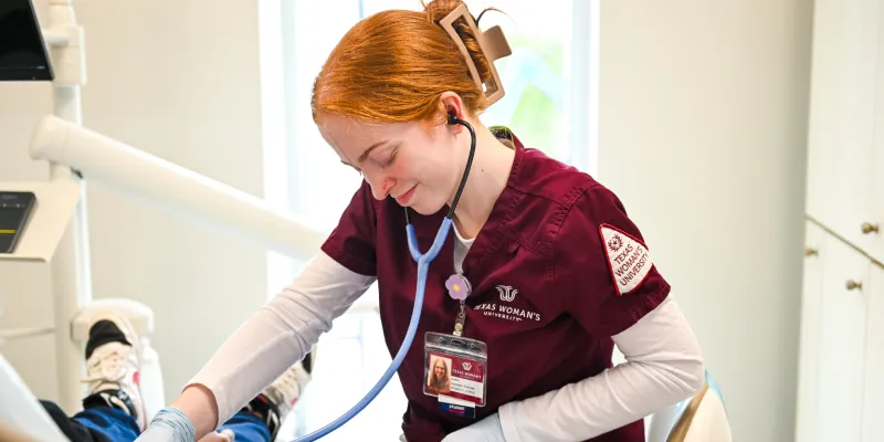 A nursing student in TWU scrubs is smiling while seated and looking down, using a stethoscope on a patient out of view.