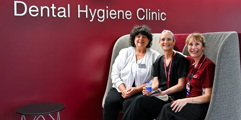 Three faculty members sit and smile in front of the Denton campus Dental Hygiene Clinic signage.