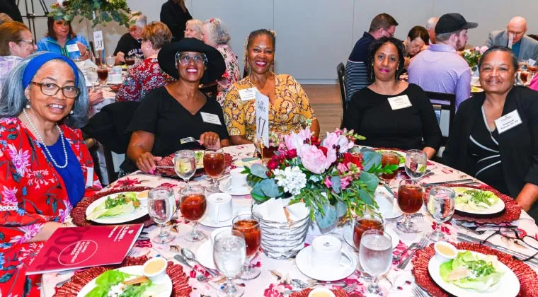 A group of smiling alumni sit around a plated meal at a table set with flowers.