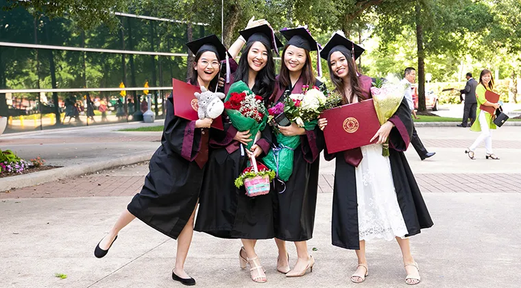 Four TWU graduates hug and smile in academic regalia.