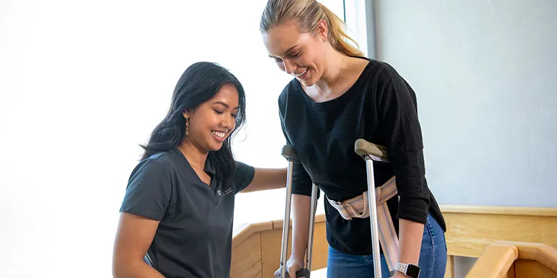 A PT student works with another student on crutches.