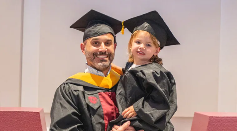 A man smiles and holds up his daughter who wears a matching cap and gown at the TWU Family Graduation ceremony.