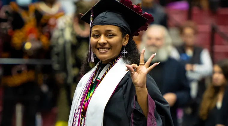 A graduate in regalia makes the TWU symbol with her left hand while smiling at the camera.