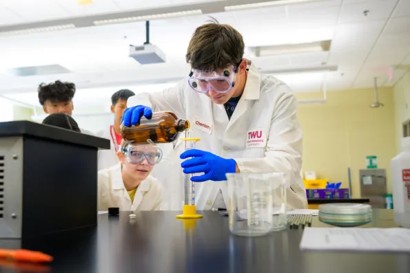 Chemistry students combine liquids in beakers in a lab setting.