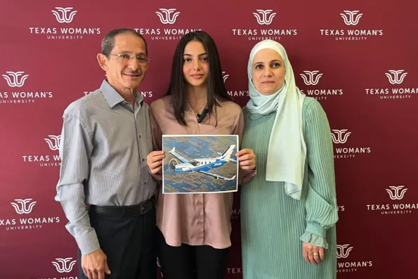 Danah Alramahi, the first recipient of the TBMOPA Foundation Scholarship, poses in front of a TWU backdrop alongside her parents. She's holding a photo of a plane and smiling.