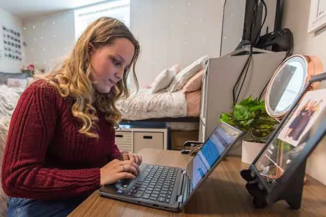 A young woman works at a laptop in her dorm room