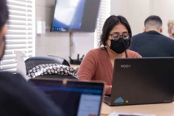 A young woman wearing a protective face mask works on a laptop with other students