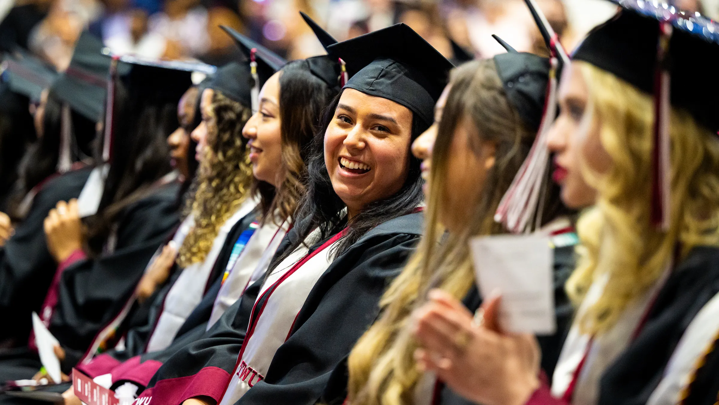 Students sit smiling in a row at commencement, the center student turned to look at the camera.