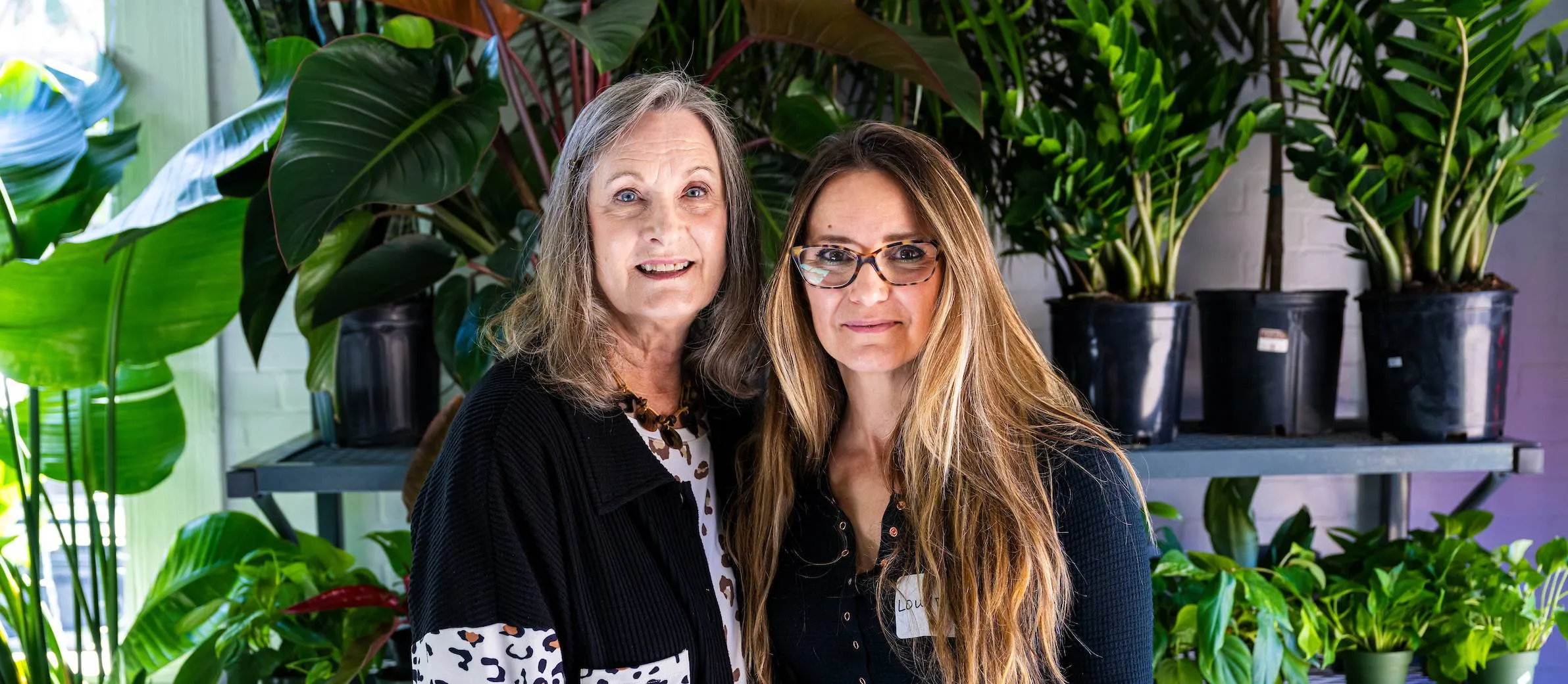 Two alumni stand together smiling at the camera in front of a lush green backdrop at True Leaf Studio in Denton.