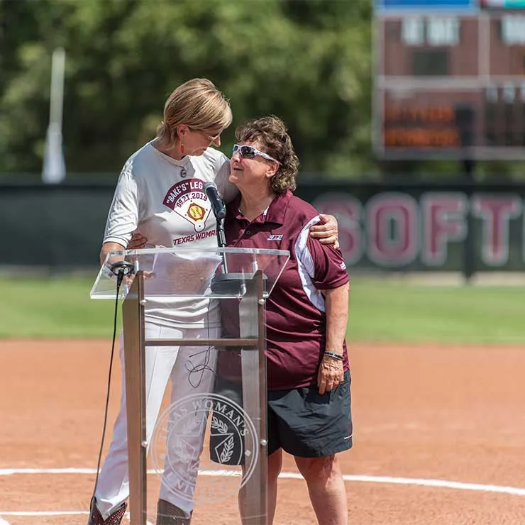 Dianne Baker and TWU Chancellor Carine Feyten hug on the new softball field on TWU's Denton campus.