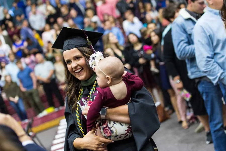 A woman walks in academic regalia during commencement while holding a baby. 