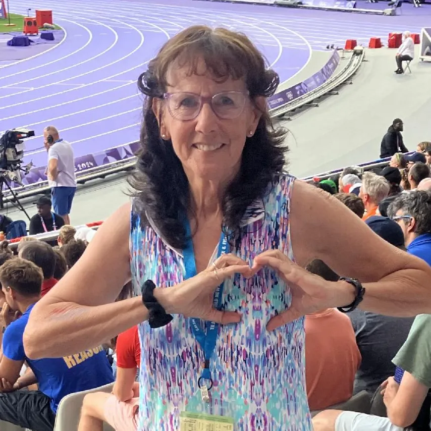 A photo of Peggy making heart hands standing in front of a running track.