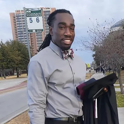 TWU graduate Ryan Matthews smiles as he stands outside the Denton campus holding his graduation cap and gown.