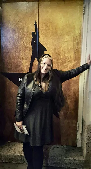 Lacy Brown stands in front of a doorway with the Hamilton Broadway poster on it