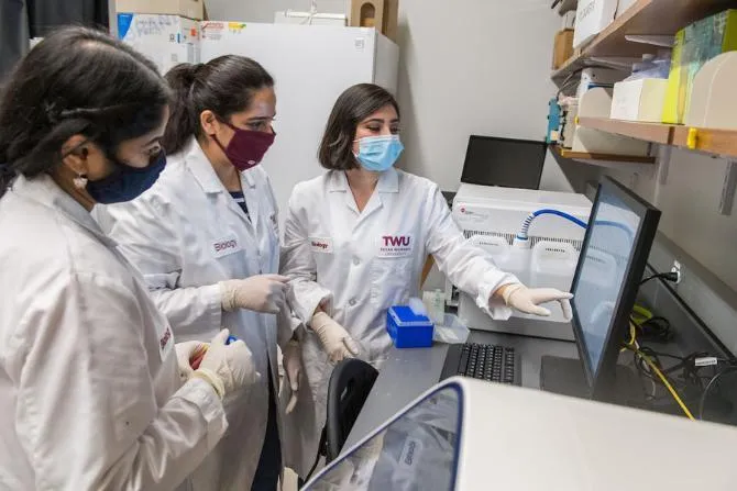 TWU Biology students in lab coats and surgical masks study specimen results on a computer screen.