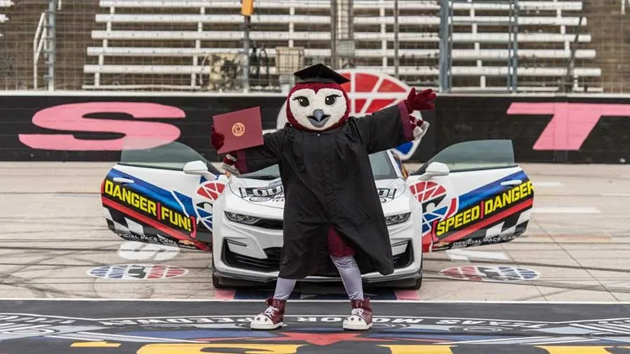 TWU mascot Oakley poses in front of a racecar at the Texas Motor Speedway, dressed in graduation regalia and holding a diploma