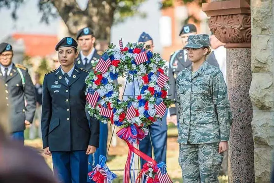 Women in military dress standing at attention with a wreath decked in American flags and ribbons