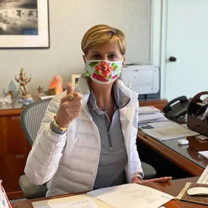 Chancellor Feyten wears a protective face mask at her office desk while giving a thumbs up signal.