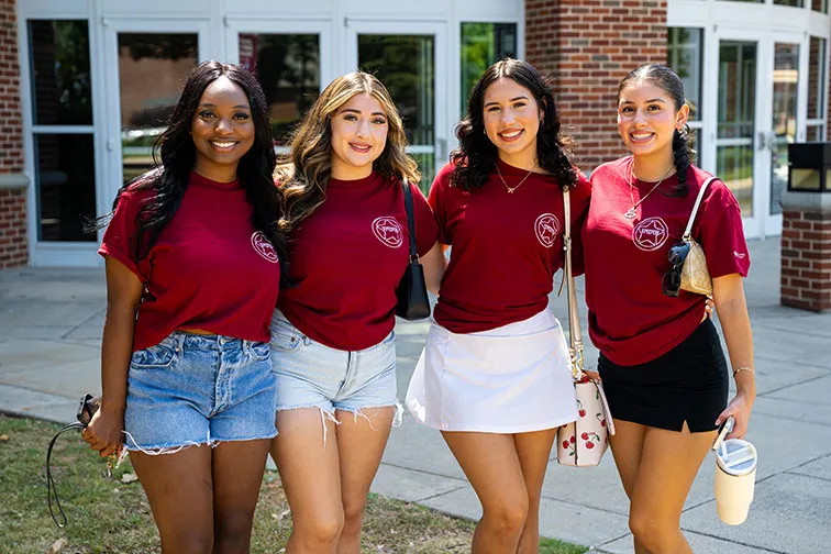 Four TWU students in maroon shirts pose for a photo outside of Pioneer Hall.