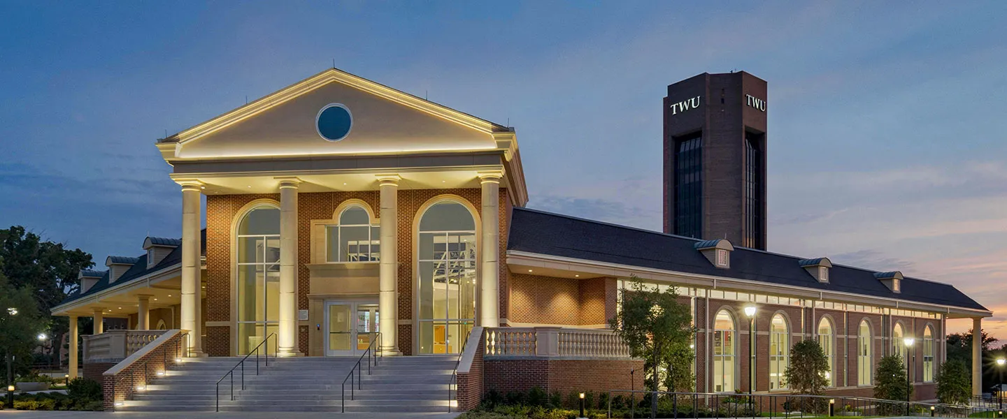 The exterior of Brackenridge Hall lit up at night with the TWU tower in the background.