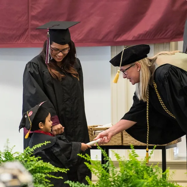 A woman hands a diploma scroll to a little boy wearing a graduation cap and gown as his mother, also in a graduation cap and gown, smiles down at him