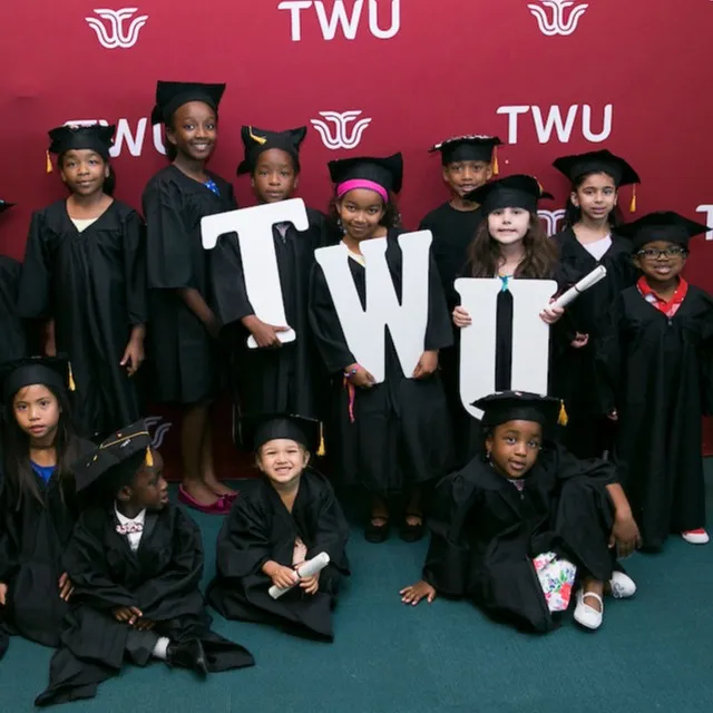 A group of kids wearing graduation caps and gowns pose around giant white letters TWU