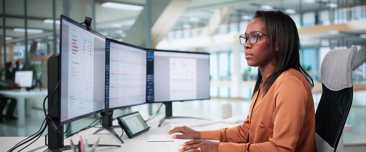 A woman in business attire works at a computer station with multiple monitors