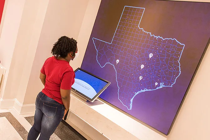 A young woman stands in front of a screen showing the districts within the state of Texas with pins marking some of the districts