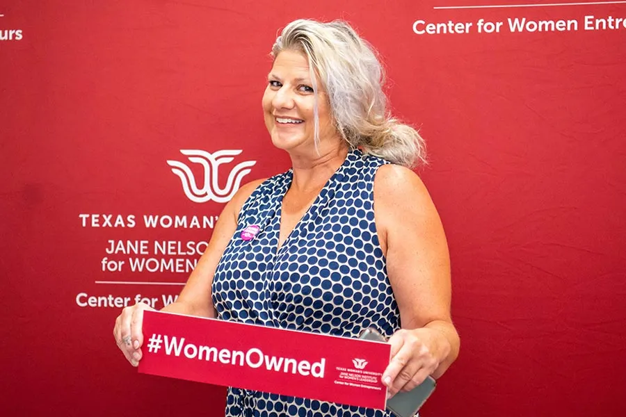 An older female entrepreneur stands holding a sign that says 'Women Owned' and the TWU logo