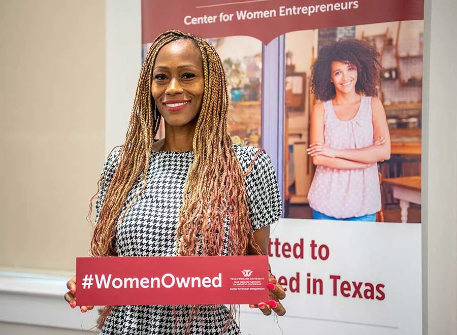 A young female entrepreneur stands holding a sign that says 'Women Owned' and the TWU logo