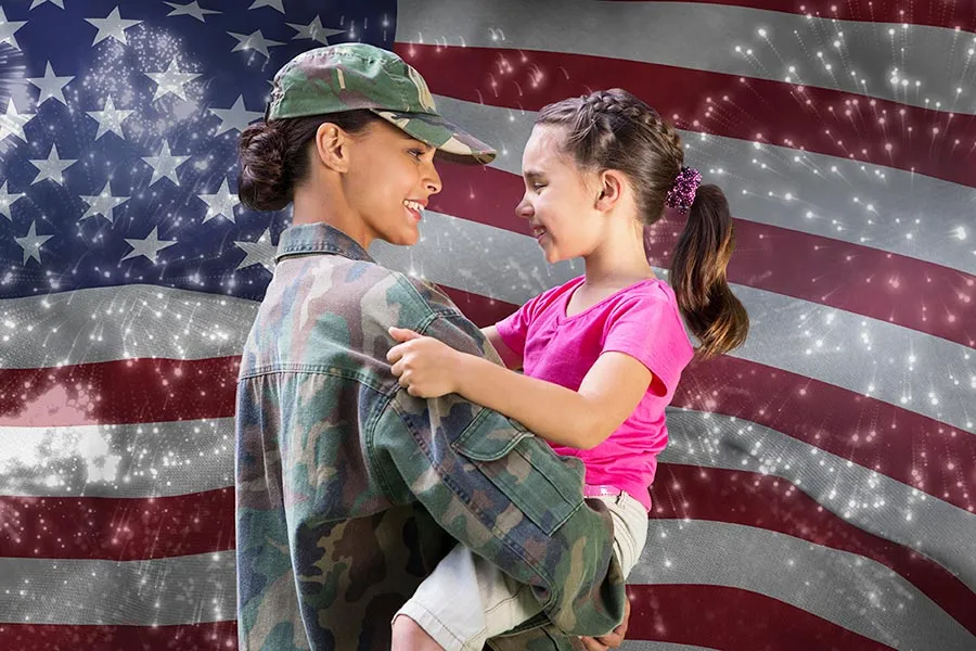 A woman in military camo holds a young child in her arms before an American flag background 