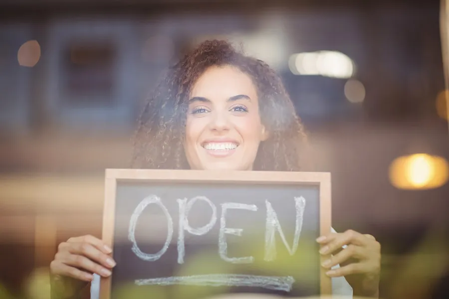 a woman standing in a shop window holding up a small blackboard sign that says 'open'
