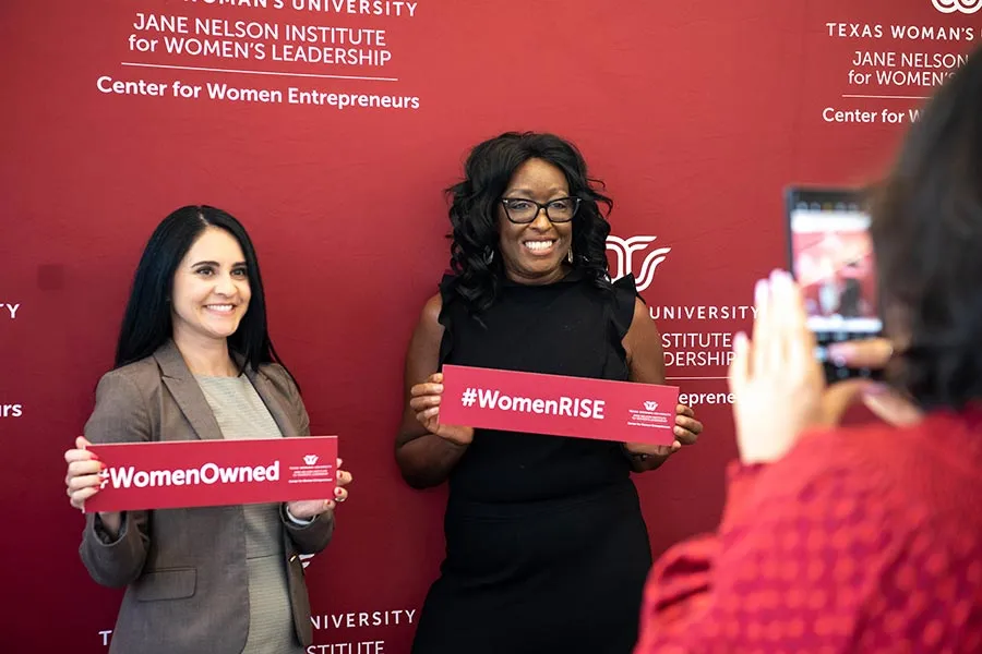 Two women entrepreneurs stand in front of a TWU Center for Women Entrepreneurs backdrop, holding signs with the Texas Woman's logo that say 'Women Owned' and 'Women RISE'