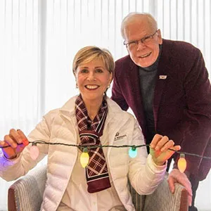 Chancellor Feyten and husband Ambassador Chad Wick smile together holding a string of colorful Christmas lights