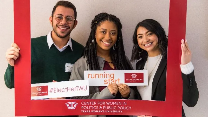 From left to right: Myah Anderson, TWU SGA president; Muhammad Kara, UNT SGA president; Kyra Solis, TWU Health Studies major; posing inside a TWU Center for Women in Politics & Public Policy frame and holding signs which read ‘Texas Woman’s University: Elect Her TWU’ and ‘Texas Woman’s University: running start’.