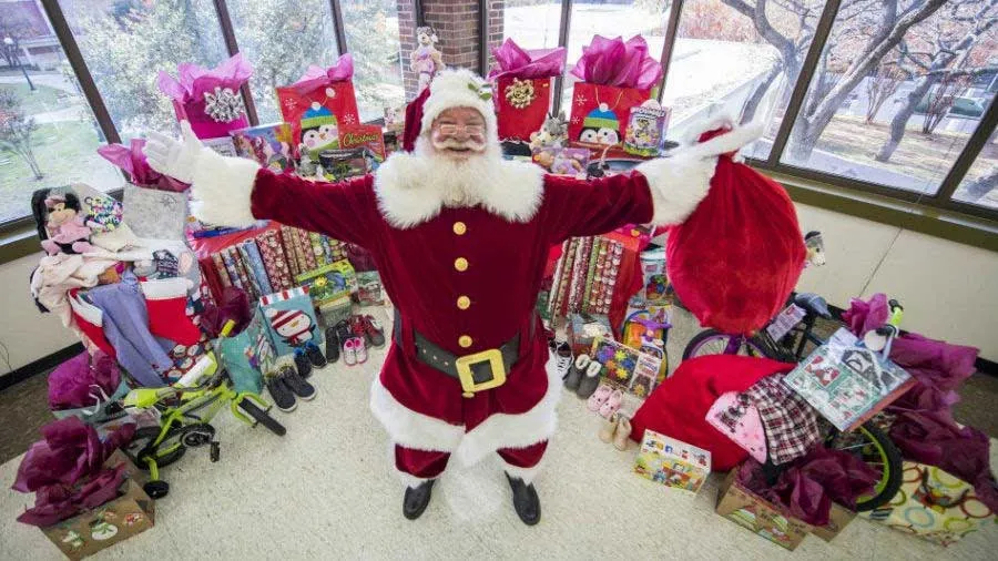 Santa Claus stands with arms outstretched with piles of gifts surrounding him