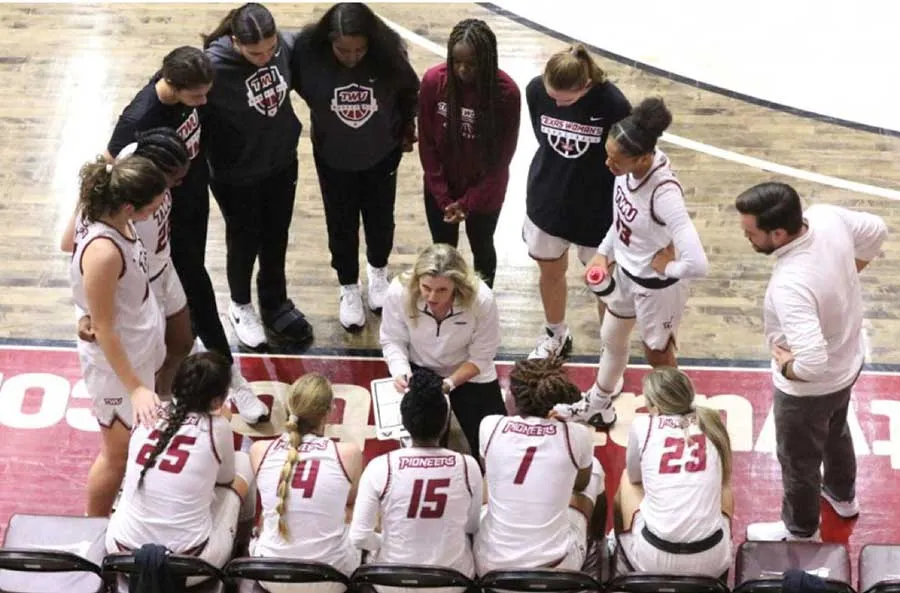 The TWU basketball team in a huddle with their coach on the court sidelines