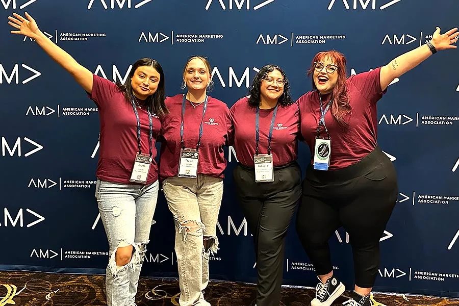Four young women dressed in maroon polos stand in front of a AMA blue backdrop