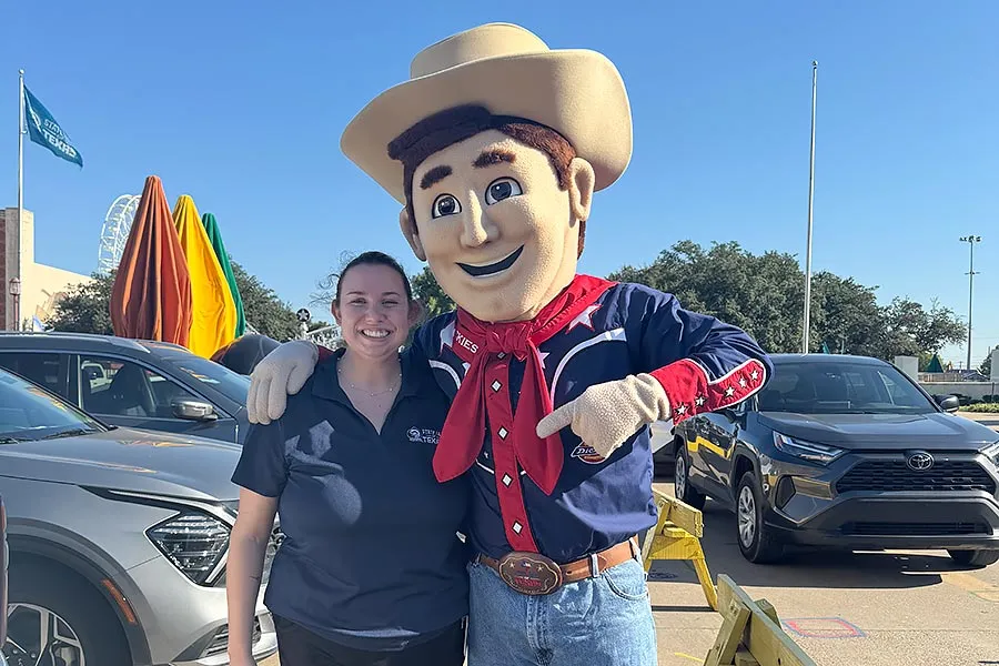 a èƵ student in a blue State Fair polo stands next to a person dressed as Big Tex