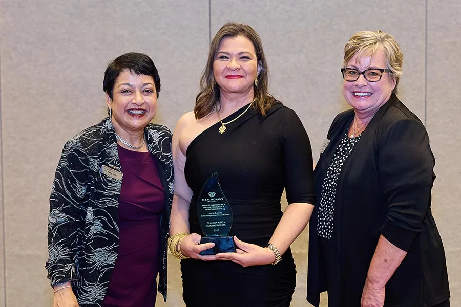 Claudia Mirza stands in the middle of three people in a black dress holding an award