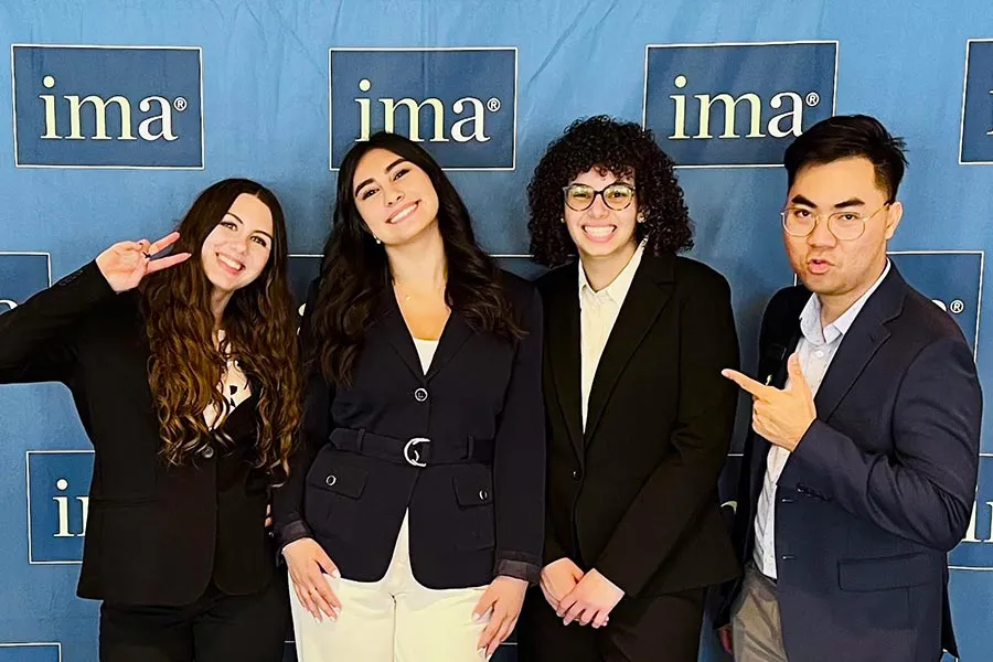 three MAK College students stand casually in business attire in front of a IMA blue backdrop along with faculty advisor