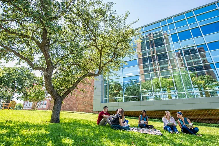 six college students sit on grass near a tree and college building