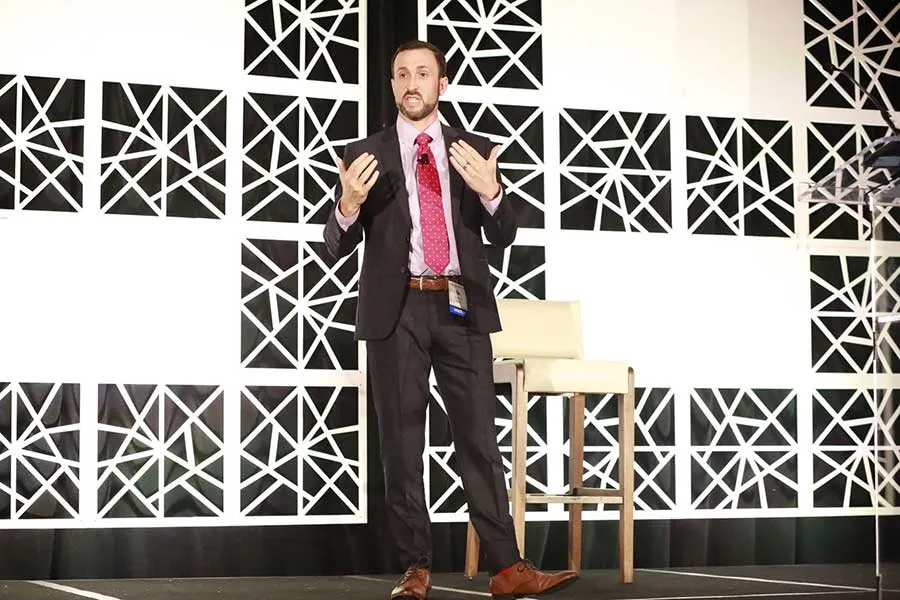 man in black suit with red tie stands on stage in front of black and shite backdrop